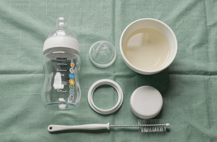 Vue à plat des composants désassemblés d'un biberon Philips Avent Natural avec accessoires de nettoyage sur tissu lin vert sauge
